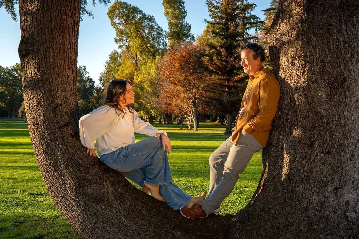 A woman and a man smiling and looking at each other while sitting against a large tree.